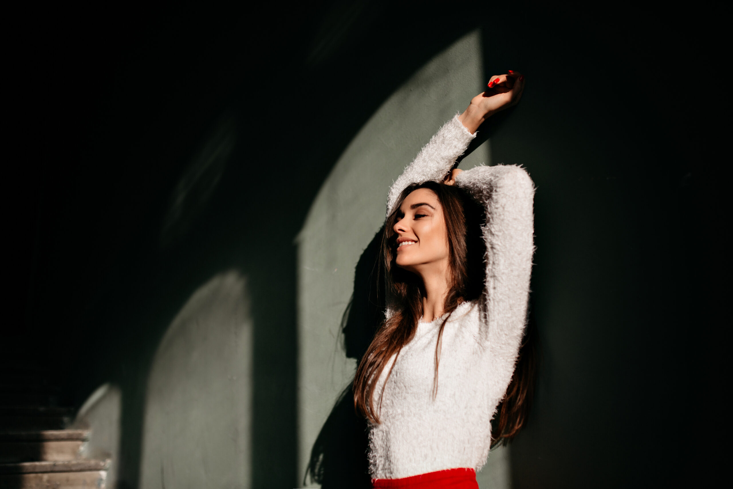 Blissful young female model with long hair standing near wall. Indoor photo of positive caucasian lady smiling in dark room..